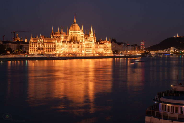 Das ungarische Parlamentsgebäude in Budapest, nachts hell erleuchtet, spiegelt sich in warmen Goldtönen auf der ruhigen Oberfläche der Donau.
