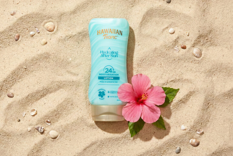 Eine türkisfarbene After Sun Lotion liegt im Sand. Daneben eine rosafarbene ne Hibiskus Blume