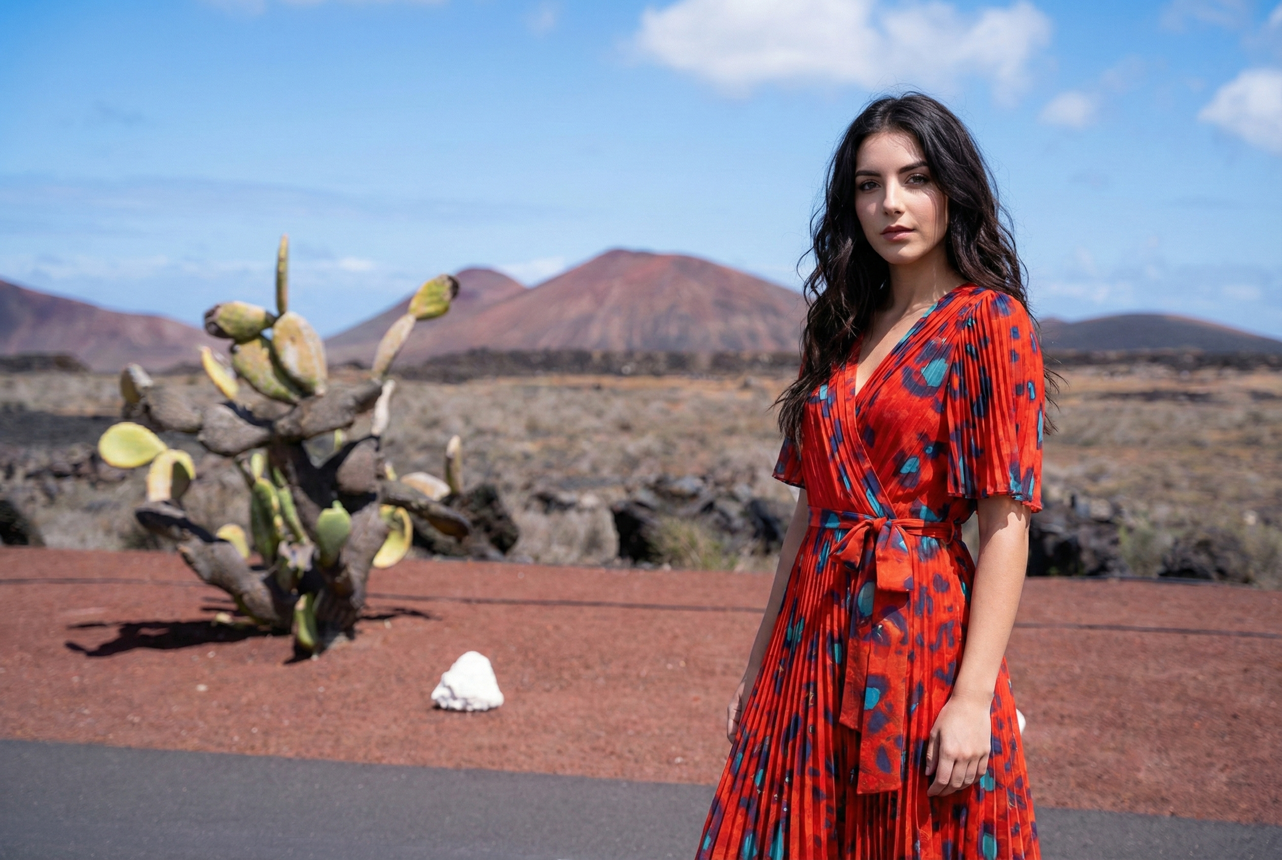 Eine junge Frau mit einem roten Kleid steht in einer Straße. im Hintergrund sind eine Vulkanlandschaft und Kakteen zu sehen.
