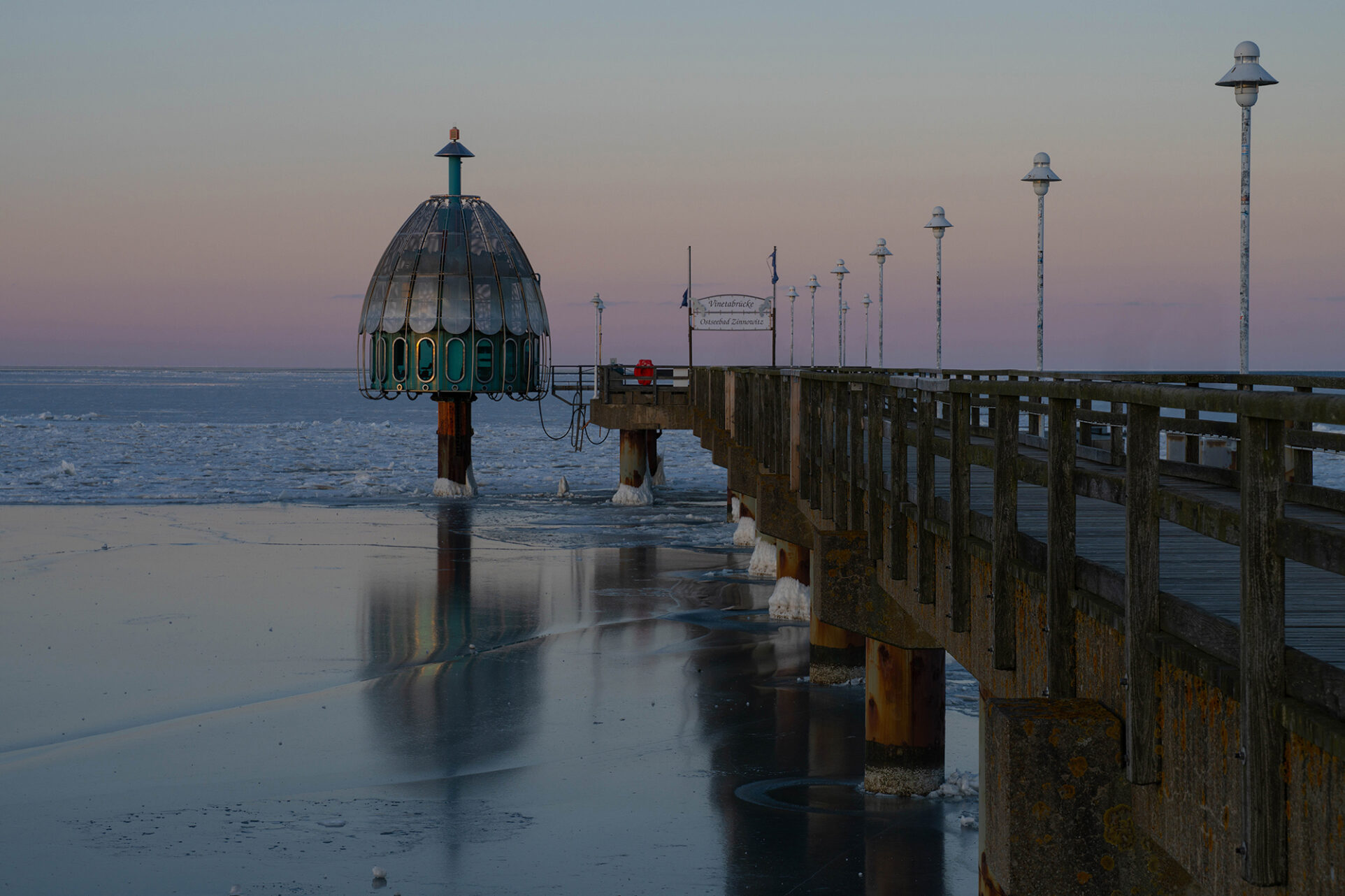 Seebrücke mit Tauchgondel im Sonnenuntergang. Die Ostsee ist gefroren und die Tauchgondel spiegelt sich auf dem Eis.