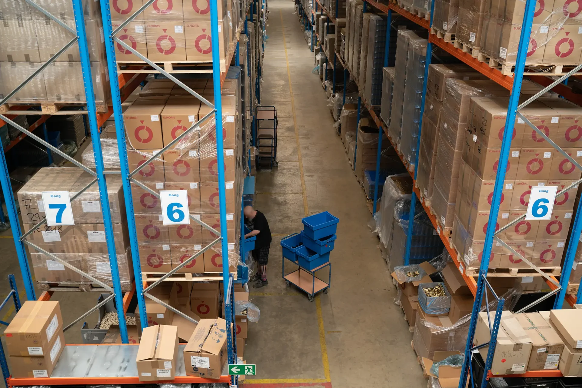 Regale in einer Lagerhalle. Ein Mitarbeiter läd Produkte auf einen Wagen.