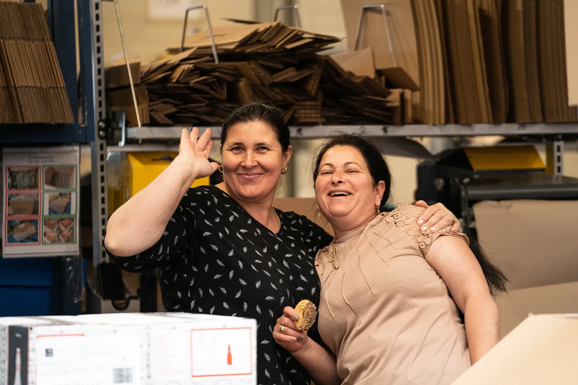 2 Mitarbeiterinnen in einer Lagerhalle winken freundlich in die Kamera.