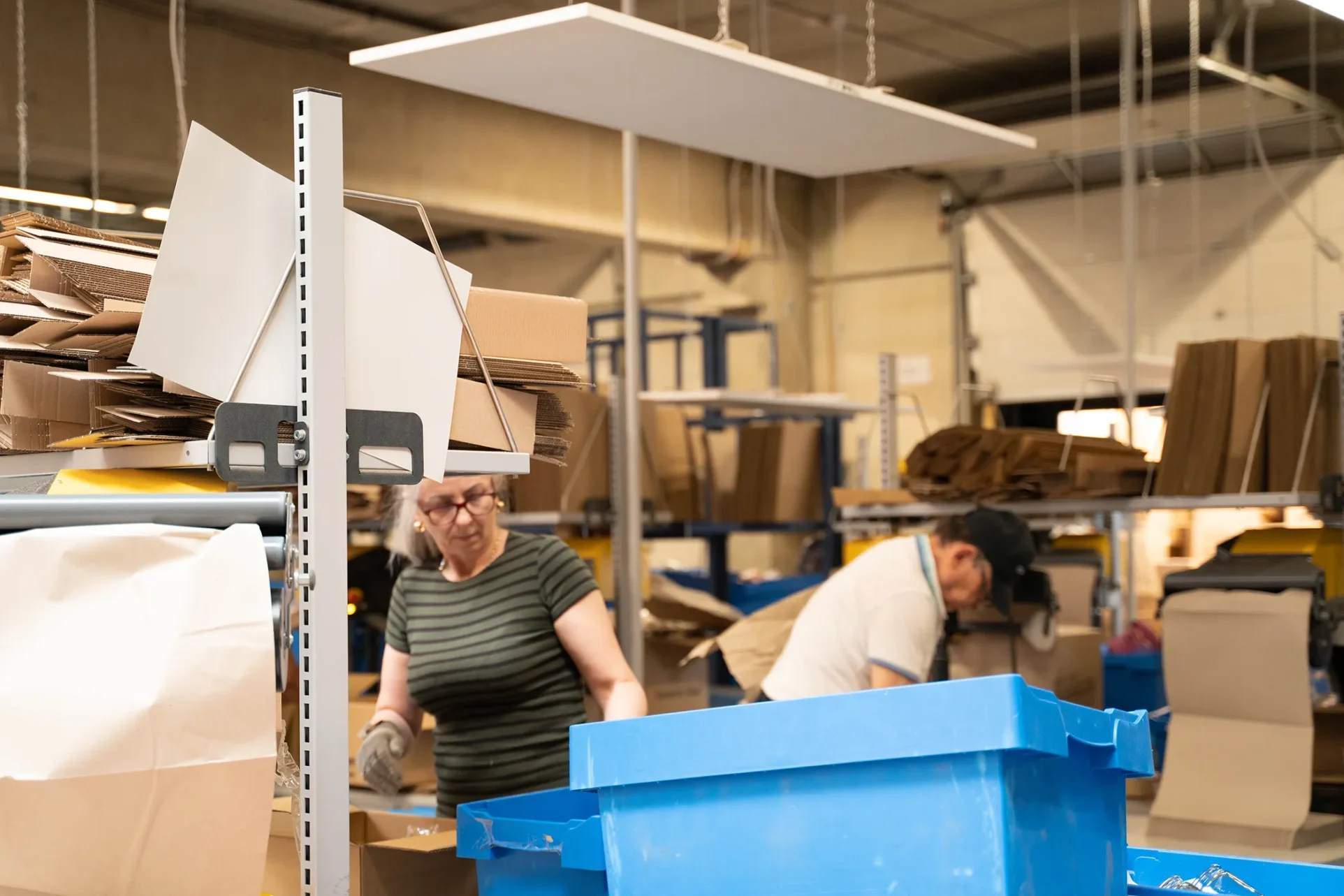 Ein Fließband mit Kisten. Im Hintergrund verpacken Mitarbeiter Artikel für den Transport.
