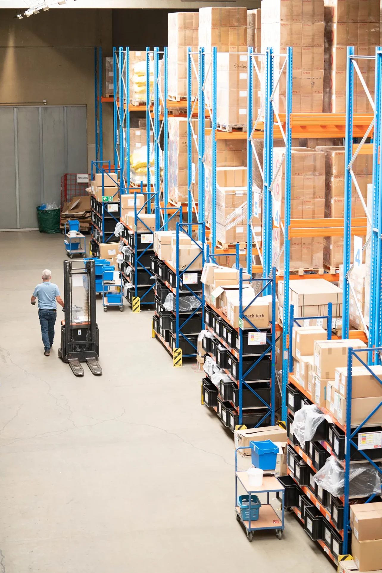 Regale in einer Lagerhalle. Ein Mitarbeiter mit einem Gabelstapler läuft an den Gegalen vorbei.
