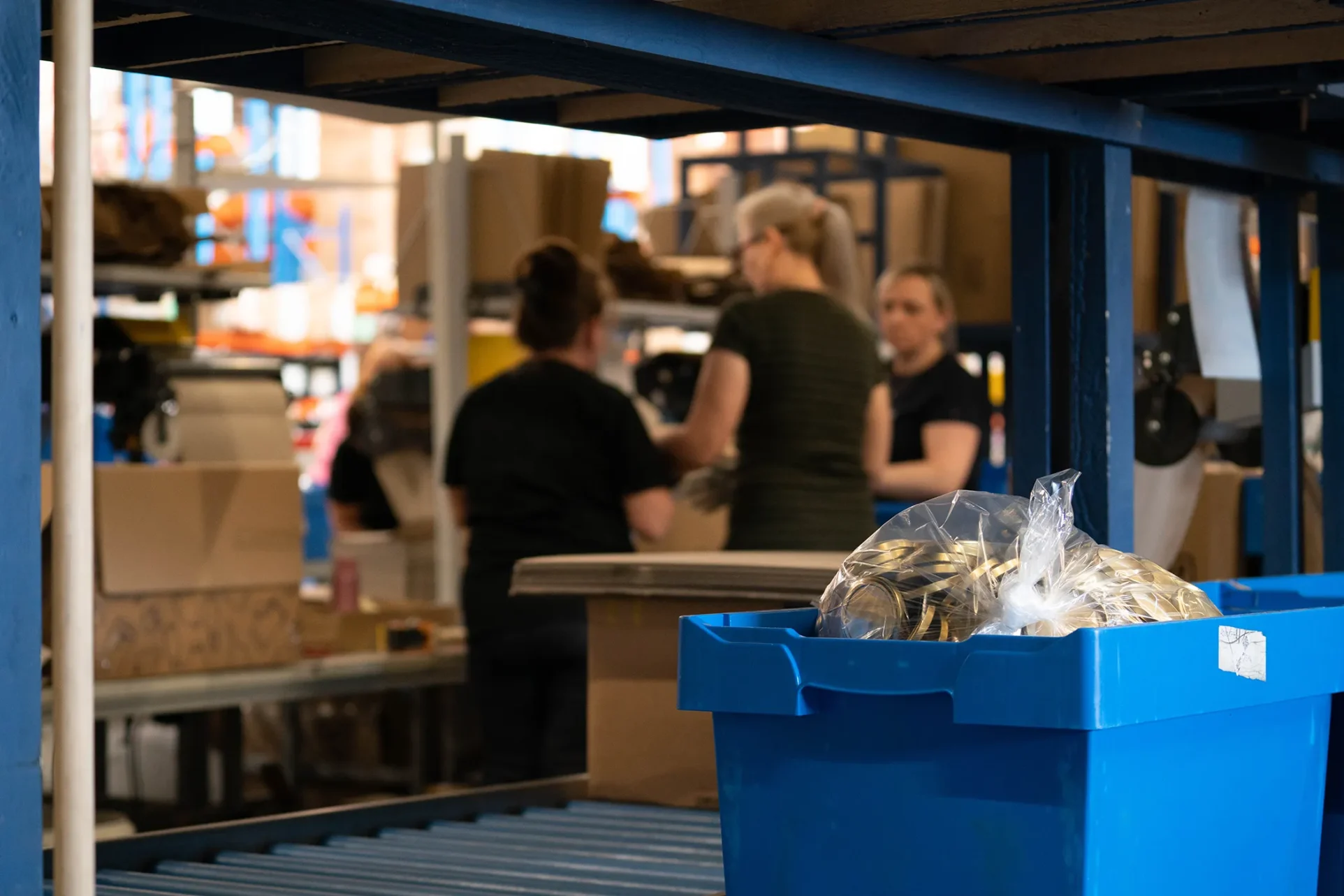 Eine Kiste mit Schraubdeckeln fährt auf einem Fließband vorbei. Im Hintergrund verpacken Mitarbeiter Produkte für den Versand.