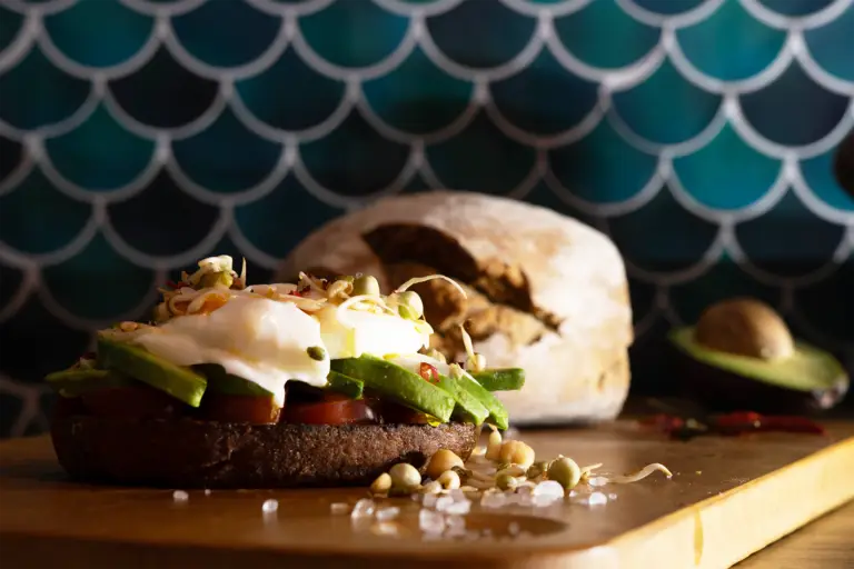 Ein Toast mit Avocado, Sprossen und Ei auf einem Holzbrett vor einem grünen Hintergrund