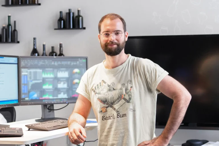 Ein Mann steht vor einem Monitor mit verschiedenen Statistiken. Er blickt in die Kamera.