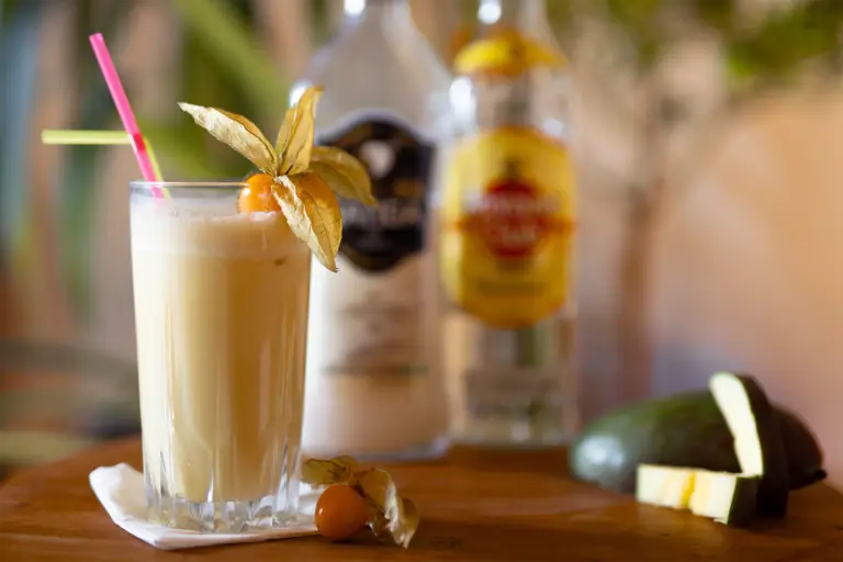 Ein glas mit einer Buttermilchgelben Flüssigkeit in einem Glas mit 2 Strohhalmen. Im Hintergrund stehen zwei Alkoholflaschen